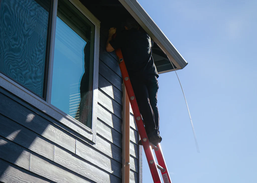 Technician working beneath a roof eave beside a window.
